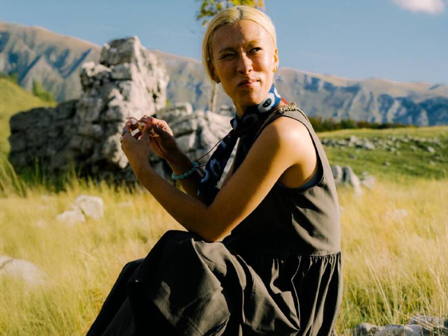 A blond woman is sitting in a green meadow, surrounded by mountains, and looking into the distance